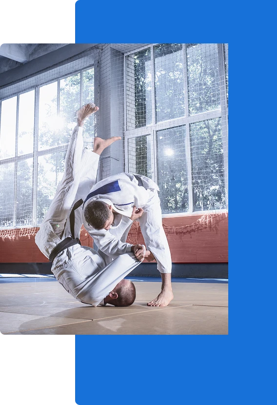 Deux pratiquants d'arts martiaux vêtus de gi blancs s'entraînent dans un gymnase bien éclairé. L'un d'eux exécute une projection de judo, faisant basculer l'autre par-dessus son épaule.